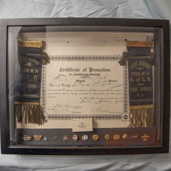 Display case with Erie Railroad Certificate of Promotion to Locomotive Engineer for Claude R. Skinner, May 20, 1924, with ribbons from Brotherhood of Locomotive Engineers Division, No. 54, Port Jervis, NY and assorted BLE anniversary pins
