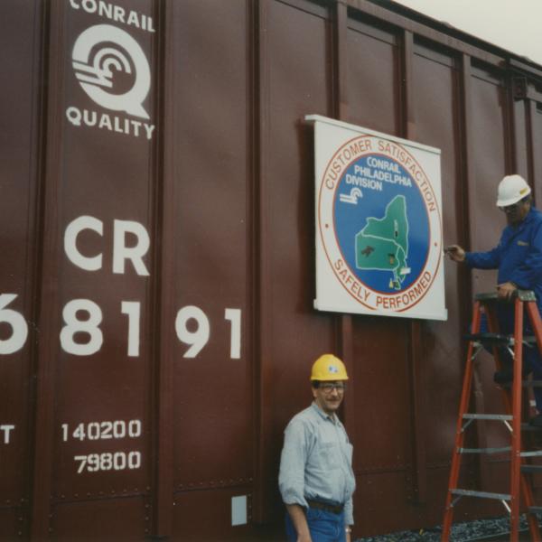 Photograph of exterior of Conrail Safety on Rails Theater boxcar 368191 with Philadelphia Division safety logo being installed, Oak Island Yard, Newark, NJ, summer 1992