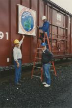 Photograph of exterior of Conrail Safety on Rails Theater boxcar 368191 with Philadelphia Division safety logo being installed, Oak Island Yard, Newark, NJ, summer 1992